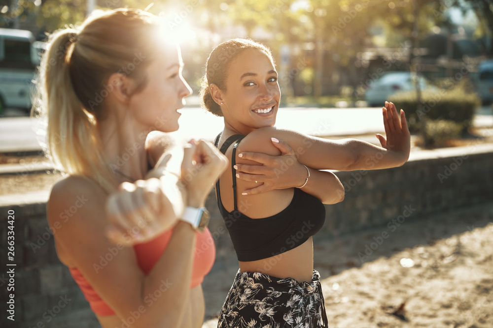 Two fit women smiling and stretching before an outdoor run - obrazy, fototapety, plakaty 