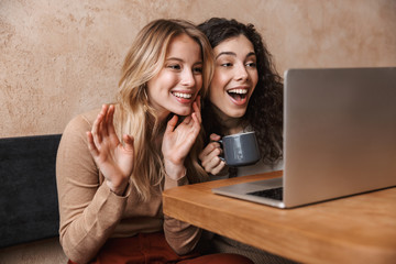 Wall Mural - Happy pretty girls friends sitting in cafe using laptop computer waving talking.