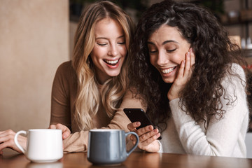 Wall Mural - Emotional shocked girls friends sitting in cafe using mobile phone.