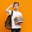 Amazed schoolboy in eyeglasses with backpack holding heap of books