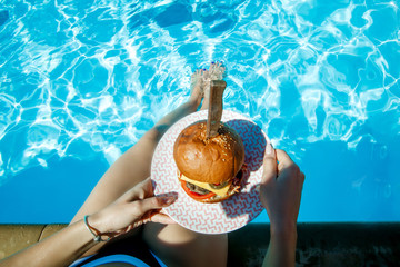 Top view of juicy Burger in girl's hands and feet in the pool. Concept burgers