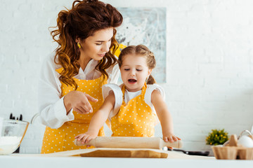 Wall Mural - daughter in polka dot apron rolling out dough on baking parchment paper while mother pointing with finger on it