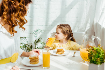 cute child drinking orange juice while having breakfast with mother