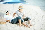 Fototapeta Natura - Happy senior couple relaxing, lying together under umbrella on the sandy beach, enjoying their retirement near the sea