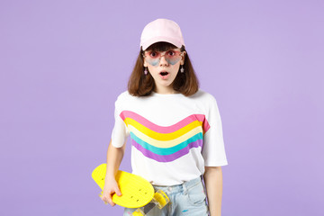 Wall Mural - Shocked teen girl in vivid clothes, heart eyeglasses holding yellow skateboard, keeping mouth open isolated on violet pastel background. People sincere emotions, lifestyle concept. Mock up copy space.