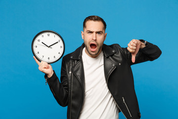 Sad upset stylish young unshaven man in black leather jacket white t-shirt holding round clock isolated on blue wall background studio portrait. People lifestyle concept. Hurry up. Mock up copy space.