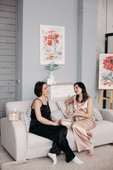 Wall Mural - Side view of two young sisters sitting on sofa at home and talking. Attractive girls in pajamas looking at each other, laughing and relaxing in morning. Concept of rest and weekends.