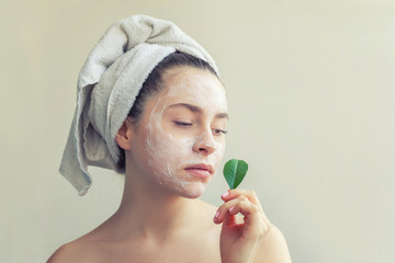 Beauty portrait of woman in towel on head with white nourishing mask or creme on face and green leaf in hand, white background isolated. Skincare cleansing eco organic cosmetic spa concept