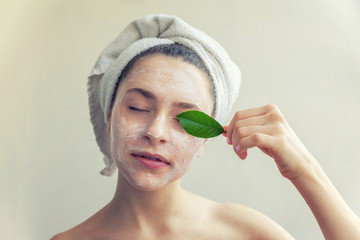 Wall Mural - Beauty portrait of woman in towel on head with white nourishing mask or creme on face and green leaf in hand, white background isolated. Skincare cleansing eco organic cosmetic spa concept