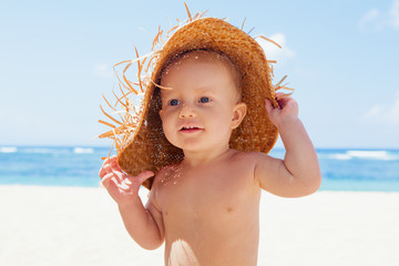 Wall Mural - On sunny white sand beach muddy happy baby boy holds hat and has fun before swimming in sea waves. Active children lifestyle, summer vacation travel with kids on tropical family resort
