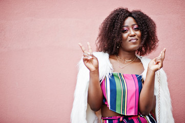 Fashionable african american woman in striped jumpsuit with fluffy faux fur coat and handbag posed against pink wall.