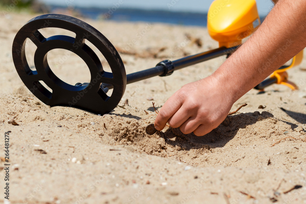 man with a metal detector on the beach picks up a coin from the sand - obrazy, fototapety, plakaty 