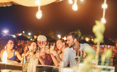 Happy friends cheering and drinking cocktails at beach party outdoor - Young millennials people having fun at weekend summer night - Youth lifestyle and nightlife concept - Main focus on right guys