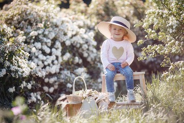 A cute little girl in a big straw hat spends time outdoors alone in a spring park park, sitting against a background of white flowering bushes. Spring portrait of a charming little girl 