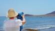 © Ana - Rear view of senior woman traveler wearing hat and holding blue telescope binocular adjusting lens to see across the sea during summer holiday on a bright sunny day in El Madano, Canary Islands, Spain