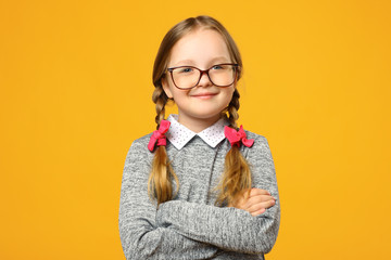 Closeup portrait of a funny little girl in glasses on a yellow background. Child schoolgirl folded her arms and looks into the camera.