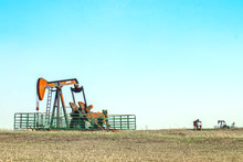 Close--up Of Oil Well Pump Jack Out In A Field Enclosed In A Metal Cattle Fence With Two Other Pumping Wells In The Distance All Set On The Horizon Of The Prairie