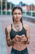 © byswat - Beautiful woman smiling, happy girl in summer in city, tanned skin tattoo. Sportswear top. Phone earphones. Concept of strength, femininity, courage, confidence, thoughts of inspiration, motivation.