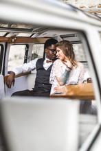 Enjoying Summer Day Togehter. Cheerful Hippie Young Couple, African Man And Caucasian Woman, Enjoying Time Together While Sitting And Hugging At Their Retro Styled Minivan. Desert Wedding