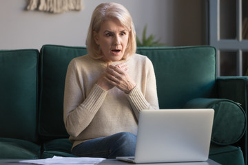 Shocked grey haired mature woman reading unexpected news on laptop