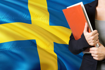 Wall Mural - Learning Swedish language concept. Young woman standing with the Sweden flag in the background. Teacher holding books, orange blank book cover.