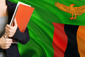 Wall Mural - Learning Zambian language concept. Young woman standing with the Zambia flag in the background. Teacher holding books, orange blank book cover.