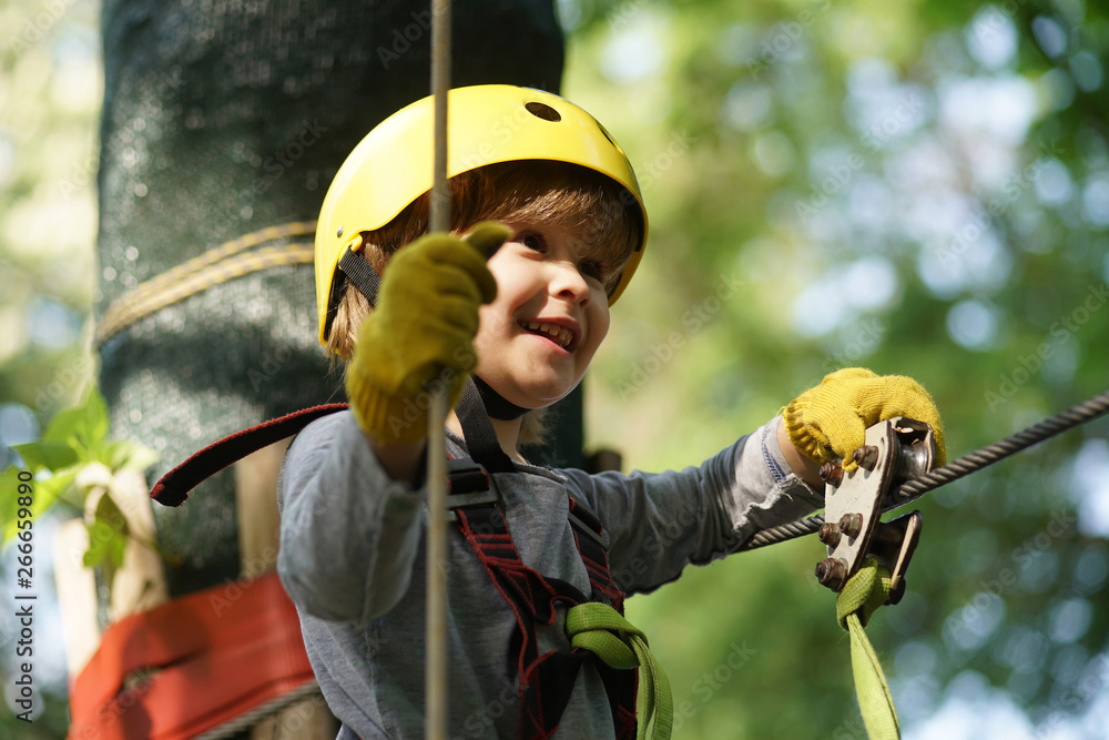 Climber child on training. Cute little boy in climbing safety equipment in a tree house or in a rope park climbs the rope. Safe Climbing extreme sport with helmet - obrazy, fototapety, plakaty 