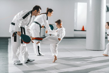 Wall Mural - Small group of kids in doboks practicing with their trainers taekwondo moves while kicking in kick target.