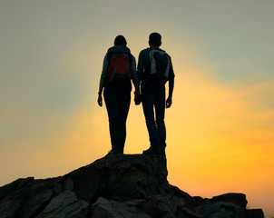 Wall Mural - couple of young travelers with backpack standing on the top of the rock at summer sunset. travel and