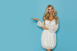 © studioloco - Beautiful Blond Woman In White Dress Is Standing With Hand Raised, Presenting And Looking At Camera