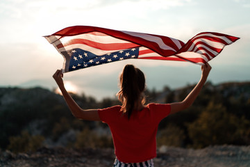 Child girl is waving American flag on top of mountain at sky background. Sunset time 