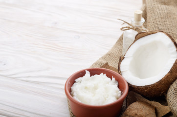 Coconut oil in brown clay bowl, glass bottle, coconut shell with meat on hemp sackcloth on white wooden kitchen table