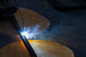 Wall Mural - Close-up view strong man is a welder in blue working overalls without gloves on arms, a metal product is welded with a welding machine in the garage workshop, blue smoke and orange sparks fly away