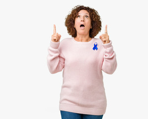 Middle ager senior woman wearing changeable blue color ribbon awareness over isolated background amazed and surprised looking up and pointing with fingers and raised arms.