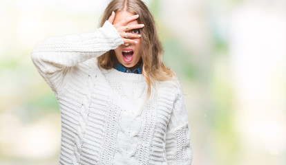 Sticker - Beautiful young blonde woman wearing winter sweater over isolated background peeking in shock covering face and eyes with hand, looking through fingers with embarrassed expression.