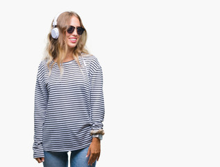 Poster - Beautiful young blonde woman wearing headphones and sunglasses over isolated background looking away to side with smile on face, natural expression. Laughing confident.