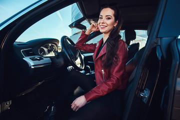 Happy young woman is sitting in her new car. She is wearing red leather jacket.