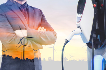 double exposure businessmen and silhouette of wind turbine at sunset with electric car battery charger station. Green energy for new innovative technology EV Electrical vehicle concept.