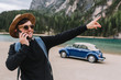 © Look! - Brunette man wears sunglasses and black sweatshirt speaking on phone during walk along river shore and pointing finger on something interesting. Outdoor portrait of laughing tourist looks on mountain.