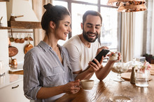 Photo Of European Brunette Couple Drinking Coffee And Using Cell Phone During Breakfast In Kitchen At Home