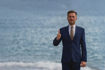 Businessman standing on the beach in a suit. Looks at camera and smiling broadly showing thumbs up gesture 