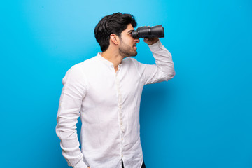 Wall Mural - Young man over isolated blue wall and looking in the distance with binoculars