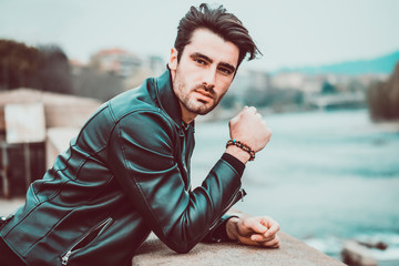 Wall Mural - One handsome young man in urban setting in modern city, standing, wearing black leather jacket and jeans, looking away