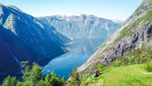 A Couple Standing On The Meadow With A Majestic View On Eidfjord From Kjeasen, Norway. Slopes Of The Mountains Are Overgrown With Lush Green Grass. Water Has Dark Blue Color. Sunny And Clear Day