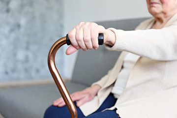 Wall Mural - Elderly woman sitting in nursing home room holding walking quad cane with wrinked hand. Old age senior lady wearing beige cardigan, metal aid stick handle bar close up. Interior background, copy space