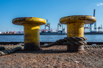 Two old Bollards on the Hamburg Harbor in Germany with rope tied around them. Harbor Cranes and Cargo Ships in the background. 