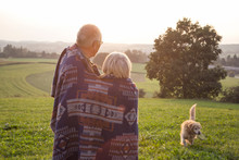 Back View Of Senior Couple Standing On A Meadow At Sunset Watching Their Dog