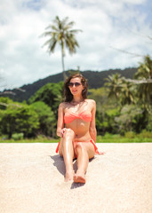 Wall Mural - Young slim woman in bikini and sunglasses sitting on fine sand beach, jungle with palm trees behind her.