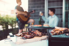 Close Up Grilled Meats And Various Food On The Grill And Celebrations Of Friends Who Are Playing Guitar And Sing Together In Their Home.