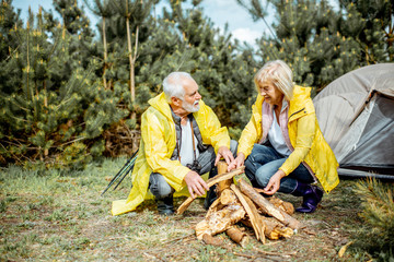 Wall Mural - Senior couple in yellow raincoats making fireplace at the campsite near the tent in the woods